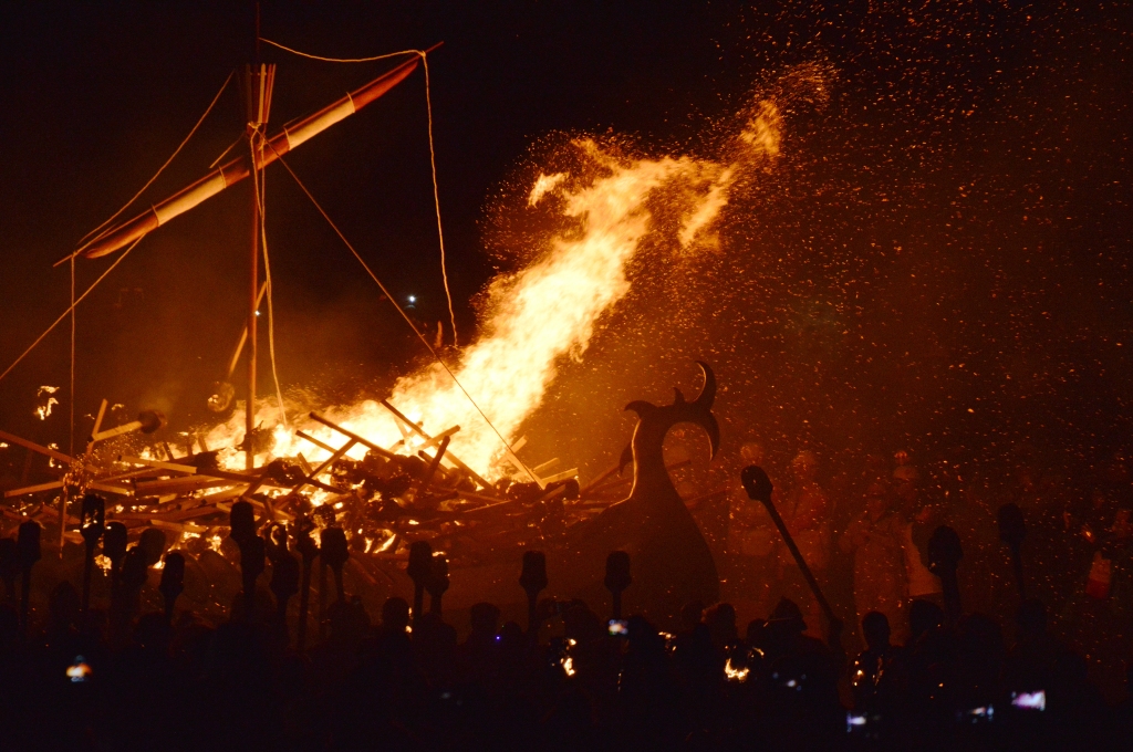 Photo at night of a Viking ship ablaze. You can see the head of the ship in a dragon shape, and the fire is only just highlighting the faces of people in costume surrounding the ship. 